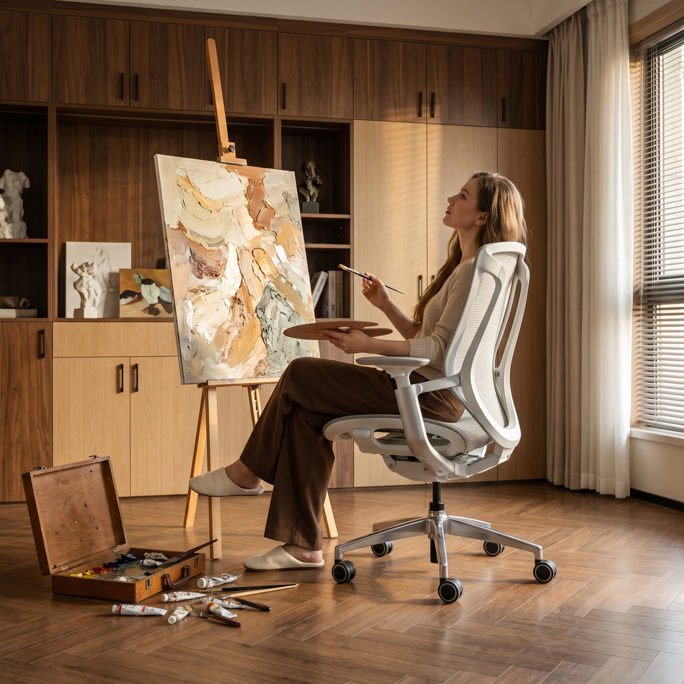 Woman painting while seated in a grey BIRCH ergonomic chair in a warm home art studio